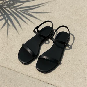 Μαύρα flat σανδάλια σε τσιμεντένια επιφάνεια με σκιά από καλοκαιρινά φυτά.